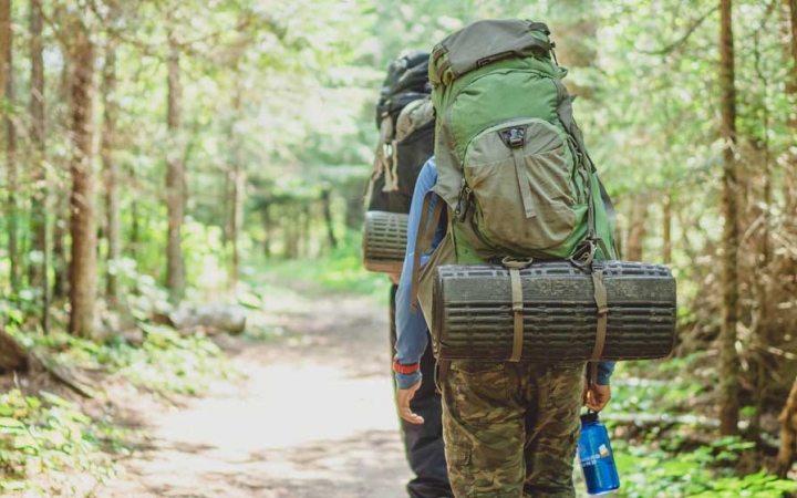 A person wearing a large backpack holds a water bottle as they hike away from the camera on a dirt trail lined with green trees. 